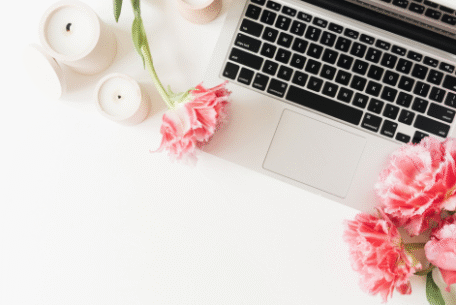 Flat lay of a laptop on a white desk surrounded by pink tulips and white candles. The flowers and candles create a soft, feminine workspace aesthetic.
